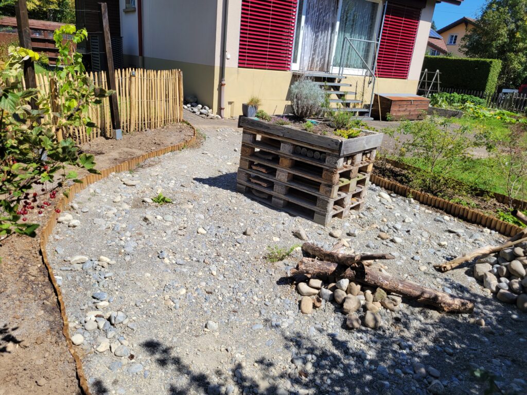fertig gestaltete Ruderalfläche: in der Mitte steht ein Insektenhotel aus Plaetten gebaut, an zwei Orten sind Steinhaufen aufgeschichtet mit grösseren Totholzästen belegt